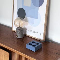 Domusnord Connector stikd�se i bl�. Kvadratisk design forl�ngerledning med 4 udtag. Vist p� en sk�nk med en bordlampe og plakat.